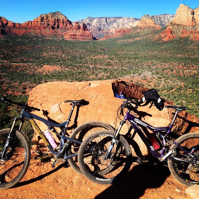Two mountain bikes are parked on a rocky outcrop overlooking a vast desert landscape with red rock formations and green vegetation. The sky is clear and blue, showcasing the natural beauty of the surrounding scenery. Hiline mountain bike trail.