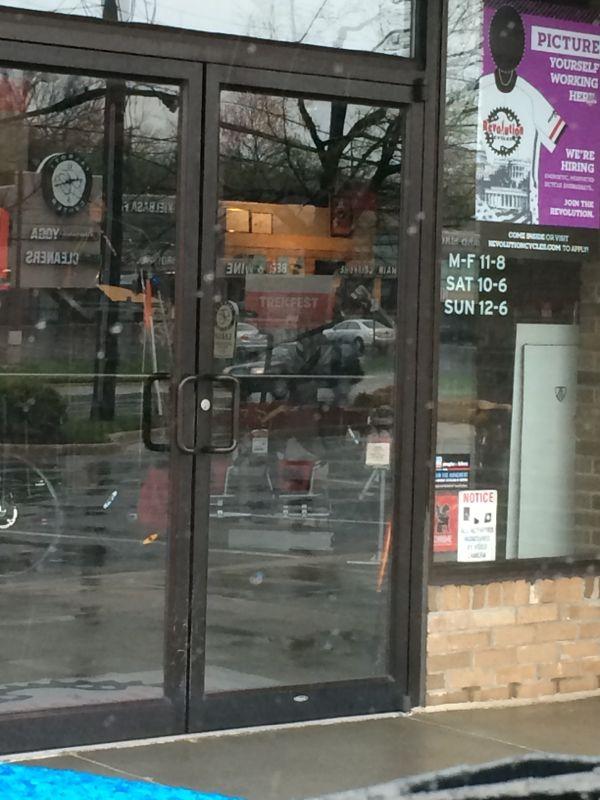 A storefront with glass double doors reflecting the surrounding area. The door features various flyers and notices, including business hours listed as Monday to Friday 11 AM to 8 PM, Saturday 10 AM to 6 PM, and Sunday 12 PM to 6 PM. A few bicycles are visible near the entrance, and the pavement appears wet, indicating recent rain.