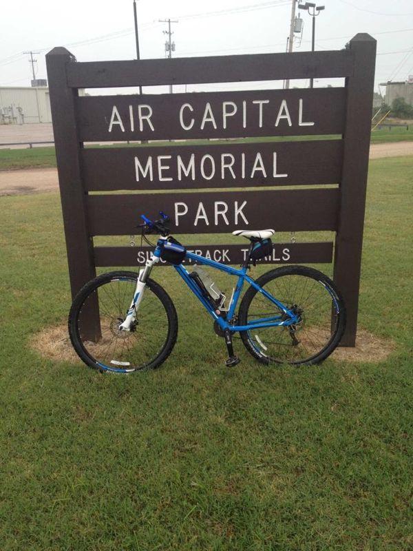 Mountain bike leaning against a wooden sign that reads "Air Capital Memorial Park" with the words "Single Track Trails" below it, set in a grassy area. Air Capital Memorial Park mountain bike trail.