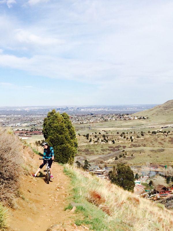 A person riding a mountain bike along a dirt trail on a hillside, surrounded by greenery and shrubs, with a view of a distant city skyline under a partly cloudy sky. Apex Park mountain bike trail.