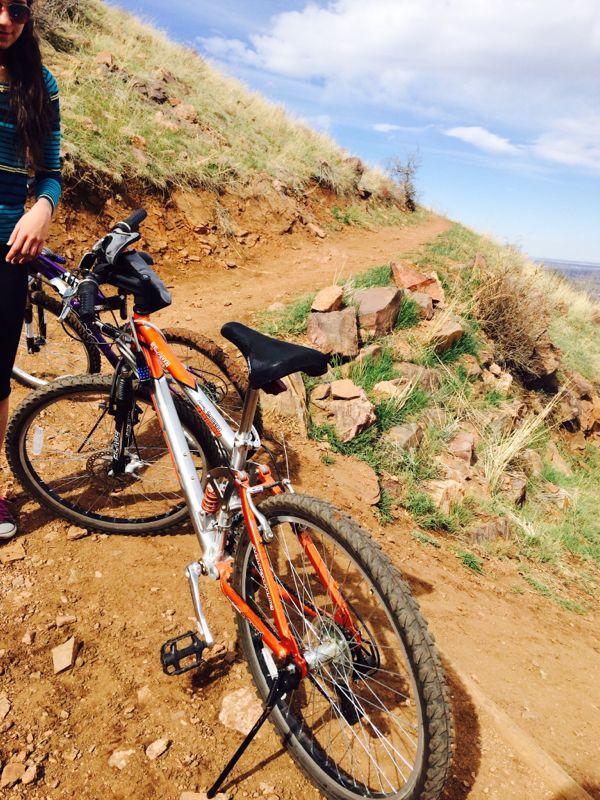 A mountain biking scene featuring two bicycles parked on a dirt trail surrounded by grass and rocky terrain. In the foreground, a person stands next to an orange and silver bike, ready to embark on their ride. The sky is partly cloudy, suggesting a bright and adventurous day for outdoor exploration. Apex Park mountain bike trail.