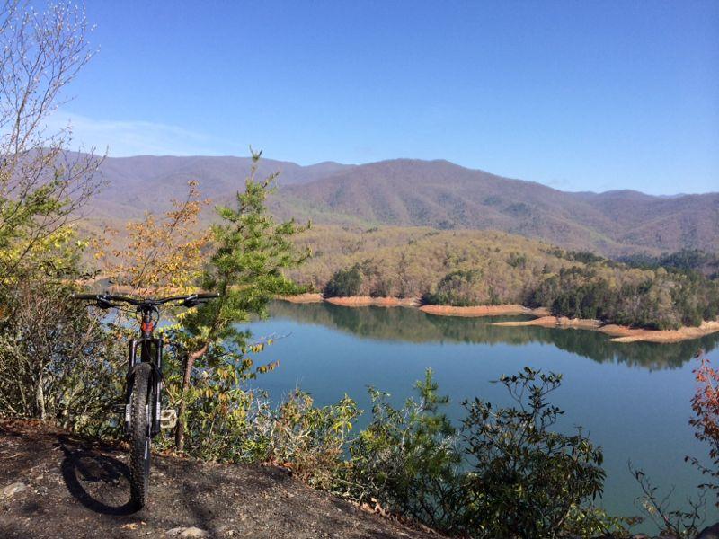 A mountain bike parked near a scenic overlook, with a tranquil lake and rolling hills in the background under a clear blue sky. Bright green trees and spring foliage frame the view, creating a peaceful nature scene. Tsali Left Loop mountain bike trail.