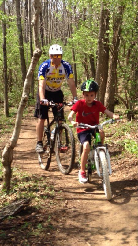 A man and young boy biking on a narrow dirt trail surrounded by trees. The man, wearing a cycling jersey and helmet, rides a mountain bike, while the boy, dressed in a red shirt and shorts, rides a smaller bike with training wheels. Veterans Park mountain bike trail.