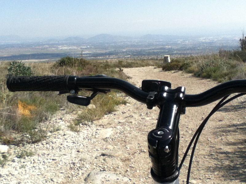 Close-up view of a mountain bike handlebar on a dirt trail, overlooking a scenic valley and distant mountains under a clear sky. The foreground features the bike