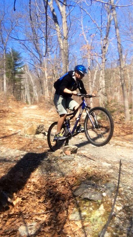 A cyclist in a blue helmet and black jacket performs a jump on a mountain bike over a rocky trail surrounded by trees and fallen leaves. The scene captures the action in a wooded area during a clear day. Lynn Woods Reservation mountain bike trail.