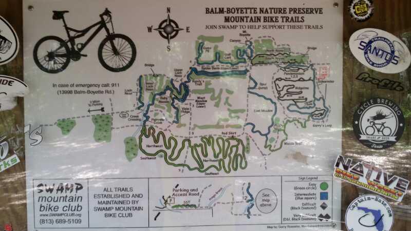 A detailed map of the Balm-Boyette Nature Preserve, showcasing mountain bike trails. The map includes labeled routes, trail difficulty levels, and important landmarks, along with a note urging visitors to call 911 in case of emergencies. There are various stickers of local biking clubs and sponsors along the edges. Balm Boyette Scrub Preserve mountain bike trail.