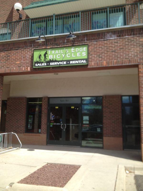 Image of the storefront for "Trail's Edge Bicycles," displaying a green and black sign that reads "Sales • Service • Rental." The shop's entrance features double doors, and there are large windows on either side. A bike rack is visible in front of the store's entrance.