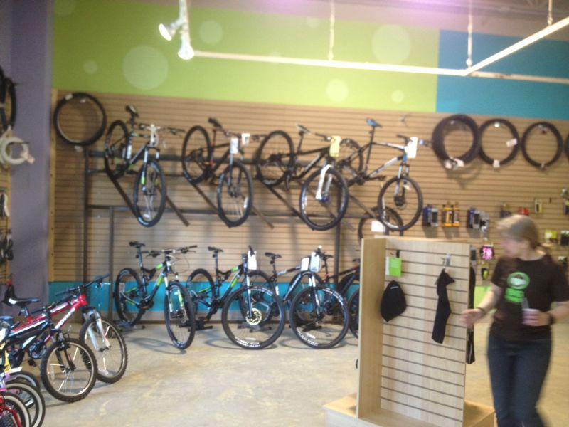 A bicycle shop interior showcasing various types of bicycles displayed on the walls, with additional bicycles arranged on the floor. The walls are painted in bright colors, and a woman is seen browsing near a display stand with accessories.
