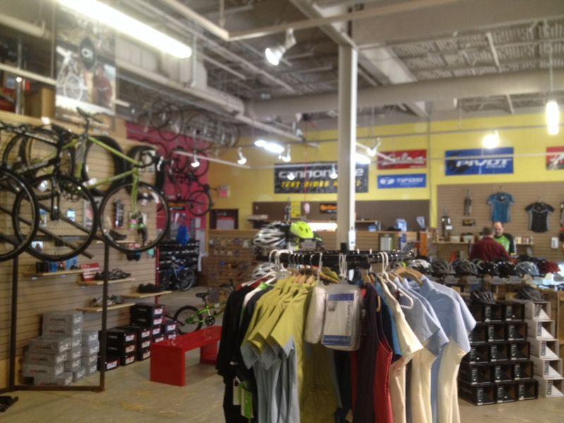 Interior of a bike shop showcasing various bicycles hanging on the walls and an organized display of cycling apparel and accessories. There are racks with t-shirts, along with helmets and other biking gear visible in the background, while customers browse the space. The shop features bright yellow walls and signage from different bike brands.