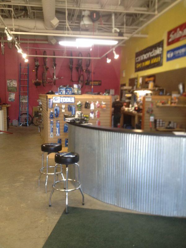 Interior view of a bicycle shop featuring a curved counter with tall stools, colorful walls, and bicycle accessories displayed on wooden panels. Tools and biking gear are visible, and bikes are hanging on the wall. The shop has a lively atmosphere with bright colors and merchandise prominently displayed.