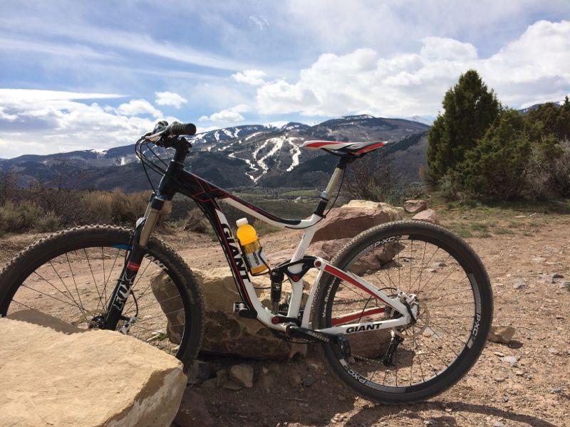 A mountain bike resting on rocks with a scenic view of mountains in the background. Snow-covered slopes are visible, and the sky features clouds, creating a picturesque outdoor setting. A water bottle can be seen mounted on the bike frame. Berry Creek Loop mountain bike trail.