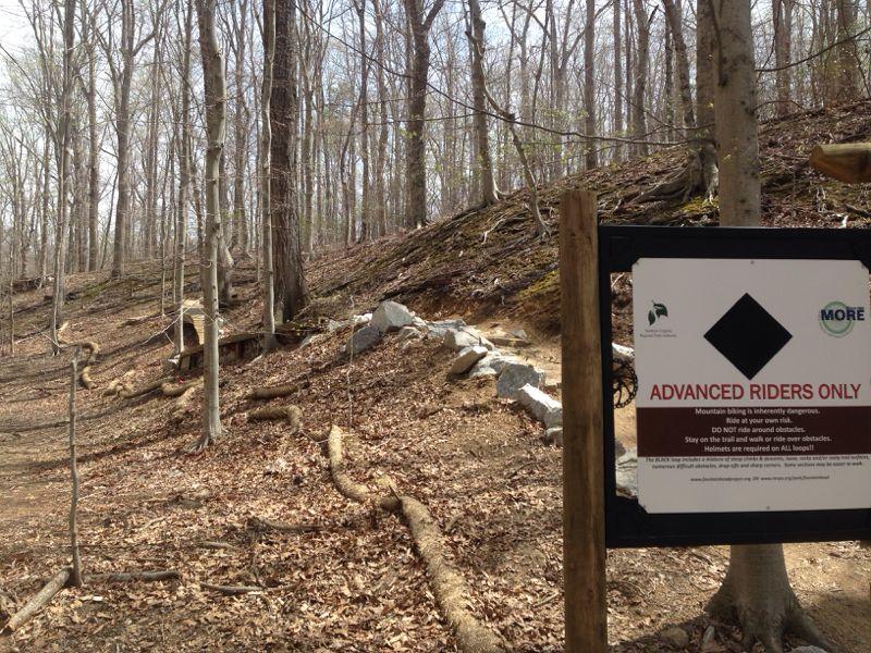 A wooded mountain biking trail with a sign that reads "ADVANCED RIDERS ONLY," warning about the dangers of mountain biking and advising riders to wear helmets. The path is rocky and surrounded by trees, with fallen leaves on the ground. Fountainhead Regional Park mountain bike trail.