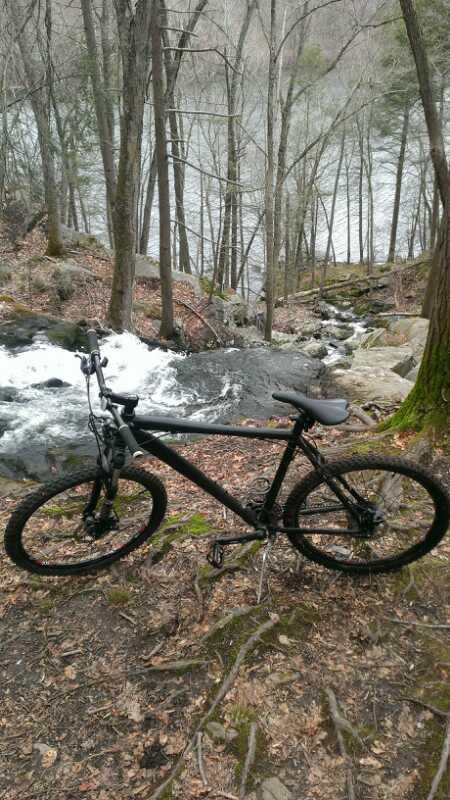 A black mountain bike positioned on a forested trail near a cascading waterfall. The bike is surrounded by bare trees and rocky terrain, with a flowing stream visible in the background. Leaves cover the ground, indicating early spring. Lower Paugussett State Forest mountain bike trail.
