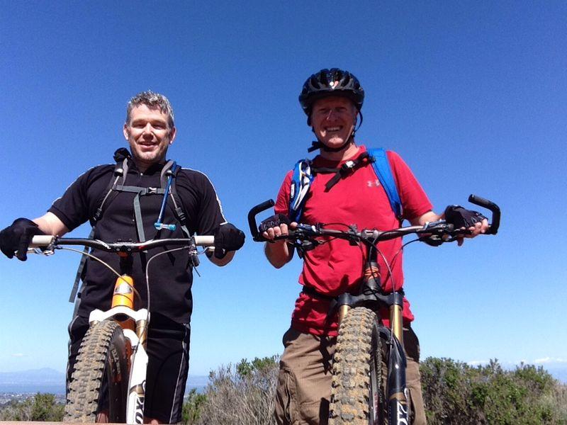 Two men smiling and posing with their mountain bikes against a clear blue sky. Both are wearing helmets and gloves, with one in a black shirt and the other in a red shirt. They appear to be enjoying a day of biking in an outdoor setting. Water Dog Lake Park mountain bike trail.