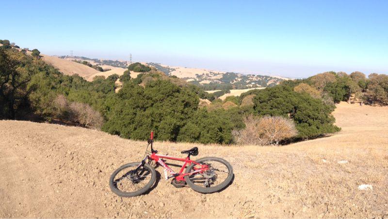 A red mountain bike resting on a dirt path, overlooking rolling hills and lush green trees under a clear blue sky. Briones Regional Park mountain bike trail.
