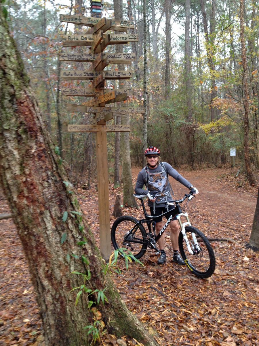 Fezzari Cascade Peak: A mountain biker wearing a helmet and casual cycling gear stands next to a wooden directional signpost in a wooded area. The ground is covered with fallen leaves, and trees surround the trail. The signpost has multiple arrows pointing in different directions, indicating various trail destinations and distances.