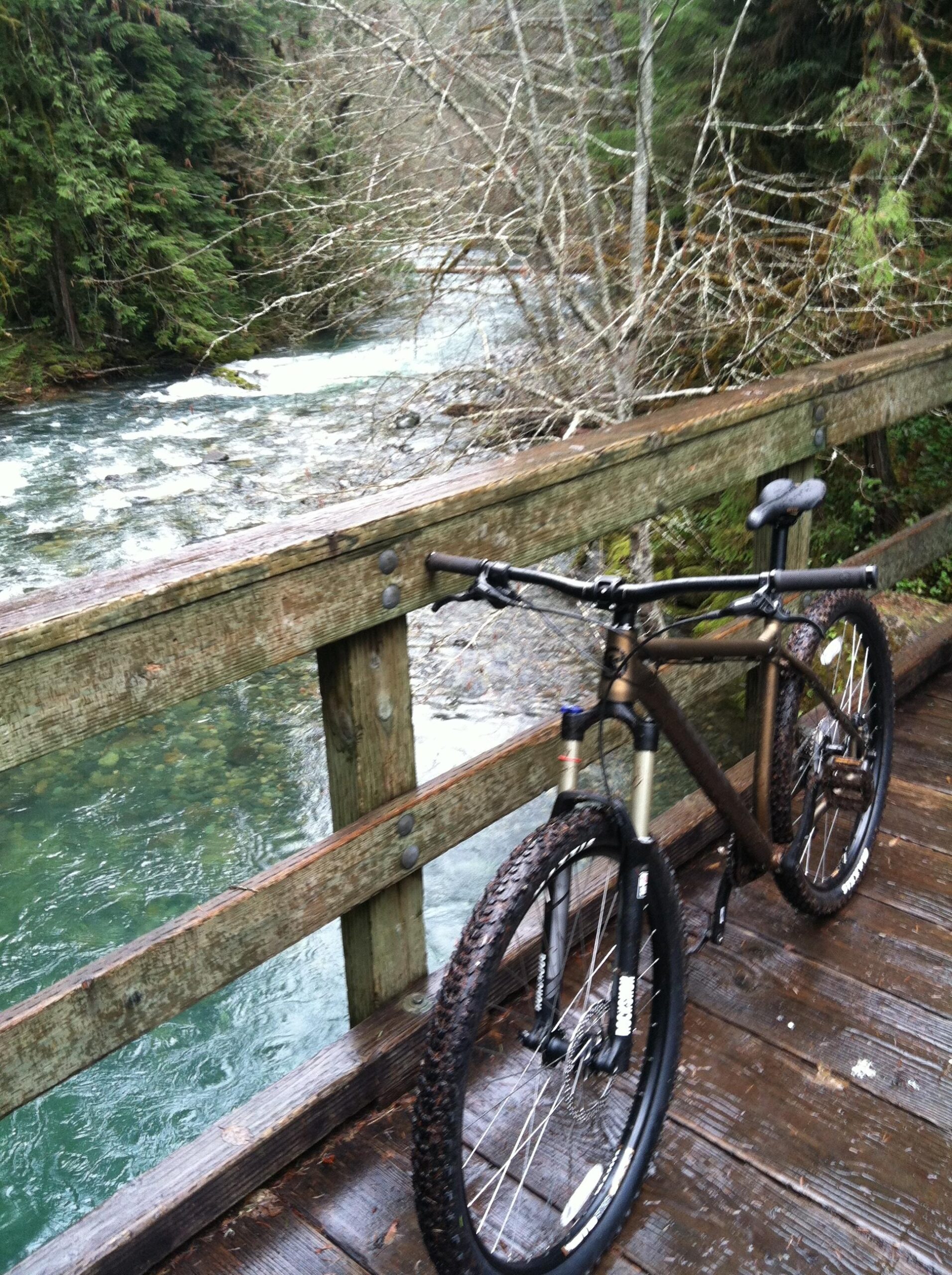 Kona Taro: A mountain bike parked on a wooden bridge overlooking a flowing river, surrounded by dense trees and a natural landscape. The scene is misty and tranquil, with wet wooden planks reflecting the calm atmosphere.