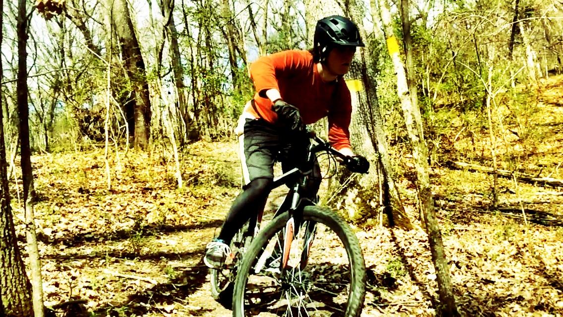 A person riding a mountain bike on a dirt trail surrounded by trees. The rider is wearing a black helmet and a red long-sleeve shirt, leaning forward as they navigate a turn on the path. The ground is covered in fallen leaves, and the atmosphere is bright and sunny. Wildwood Trails mountain bike trail.