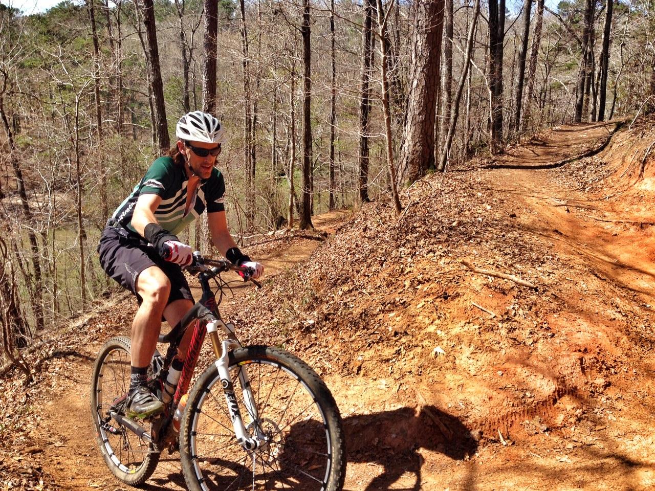 A mountain biker riding along a dirt path through a wooded area, showcasing vibrant brown soil and sparse trees in a late winter or early spring setting. The cyclist is wearing a helmet, sunglasses, and a striped jersey, demonstrating an active outdoor lifestyle. Blankets Creek mountain bike trail.