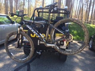 Scott Aspect Trail 29er: A mountain bike mounted on the back of a vehicle, with muddy tires, parked in a wooded area with trees in the background.