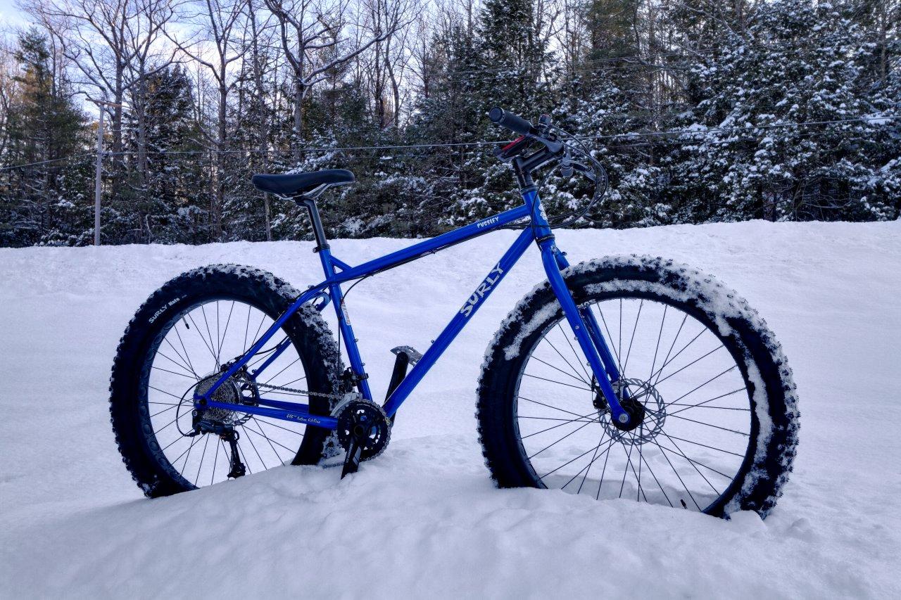 A blue fat bike parked in deep snow, showcasing its wide tires covered in snow, set against a backdrop of snowy trees and a chilly winter landscape.