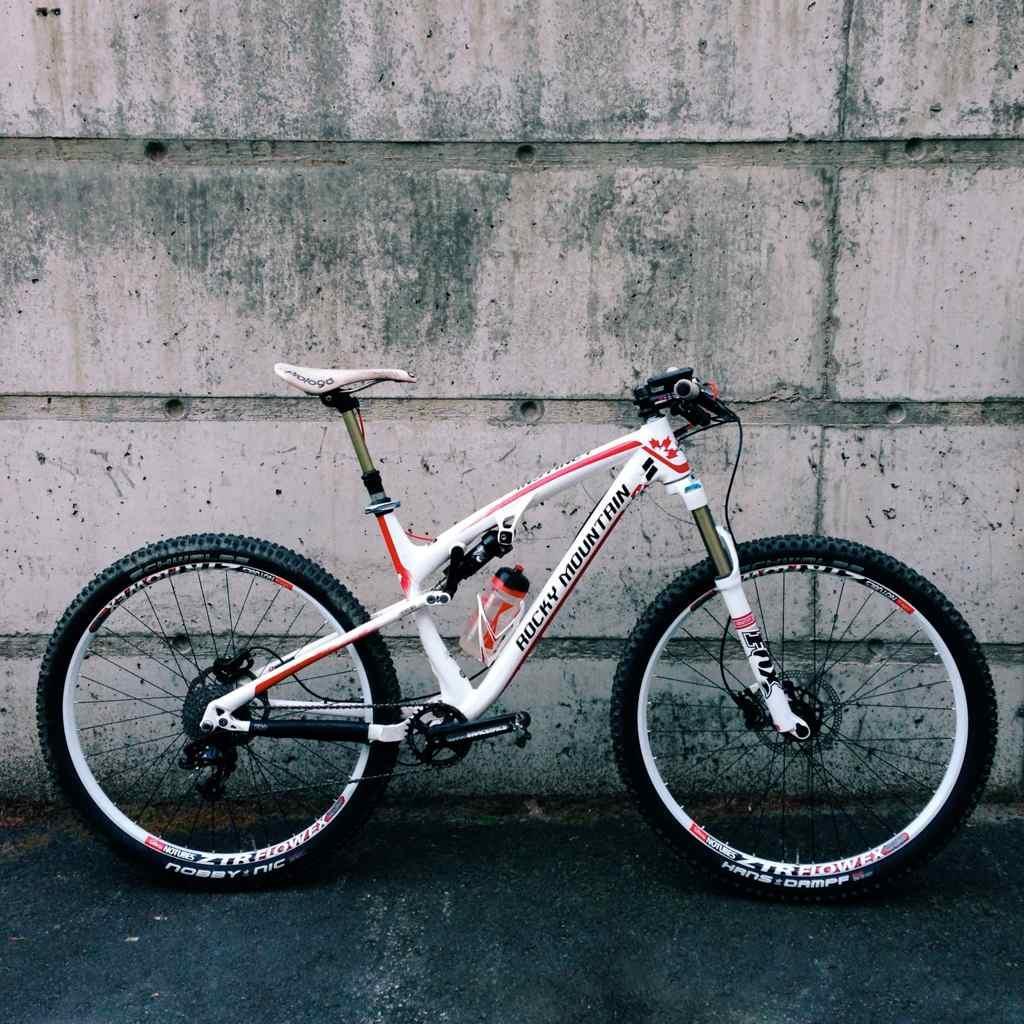 Rocky Mountain Instinct 950: A white mountain bike with red accents leaning against a textured concrete wall. The bike features wide tires and a suspension system, showcasing its design for off-road use.