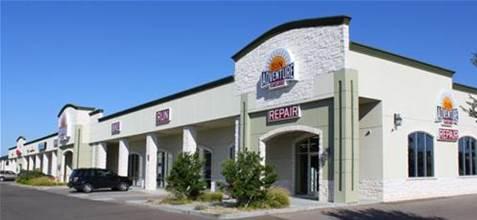 Exterior view of a commercial building with multiple storefronts, featuring signs for various businesses including "Gun" and "Repair." The building has a light-colored stone facade and large windows, set against a clear blue sky. Green landscaping is visible in front of the building.