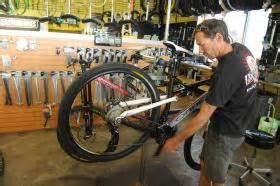 A bicycle mechanic working on a mountain bike in a bike shop, with various tools and bike parts visible on the wall behind him.