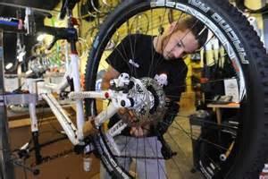 A mechanic working on a mountain bike in a bicycle repair shop, focusing on the wheel's hub and gearing system. The shop is filled with various bicycle components and tools in the background.