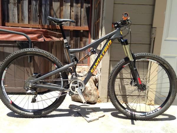 Santa Cruz Bronson: A high-quality mountain bike with a gray frame and bright yellow "Santa Cruz" branding, positioned on a flat surface. The bike features front suspension, knobby tires, and a multi-gear setup, with a wooden backdrop and a partially opened door visible in the background.