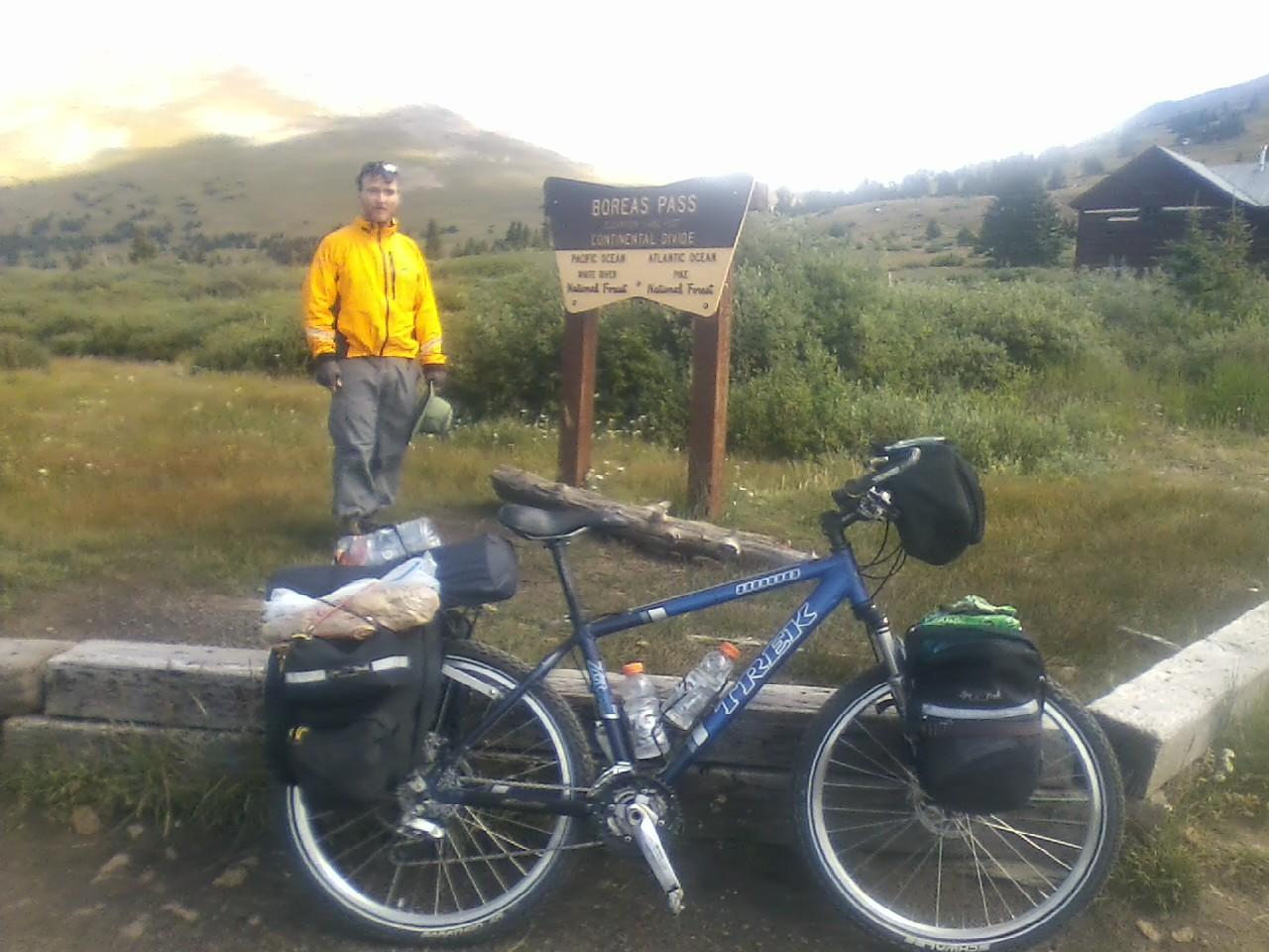 Trek 8000: A cyclist in a yellow jacket stands at the Boreas Pass sign, surrounded by mountainous landscape and greenery. Beside the bike, which is equipped with saddlebags containing supplies, scenic hills can be seen in the background under a partly cloudy sky.