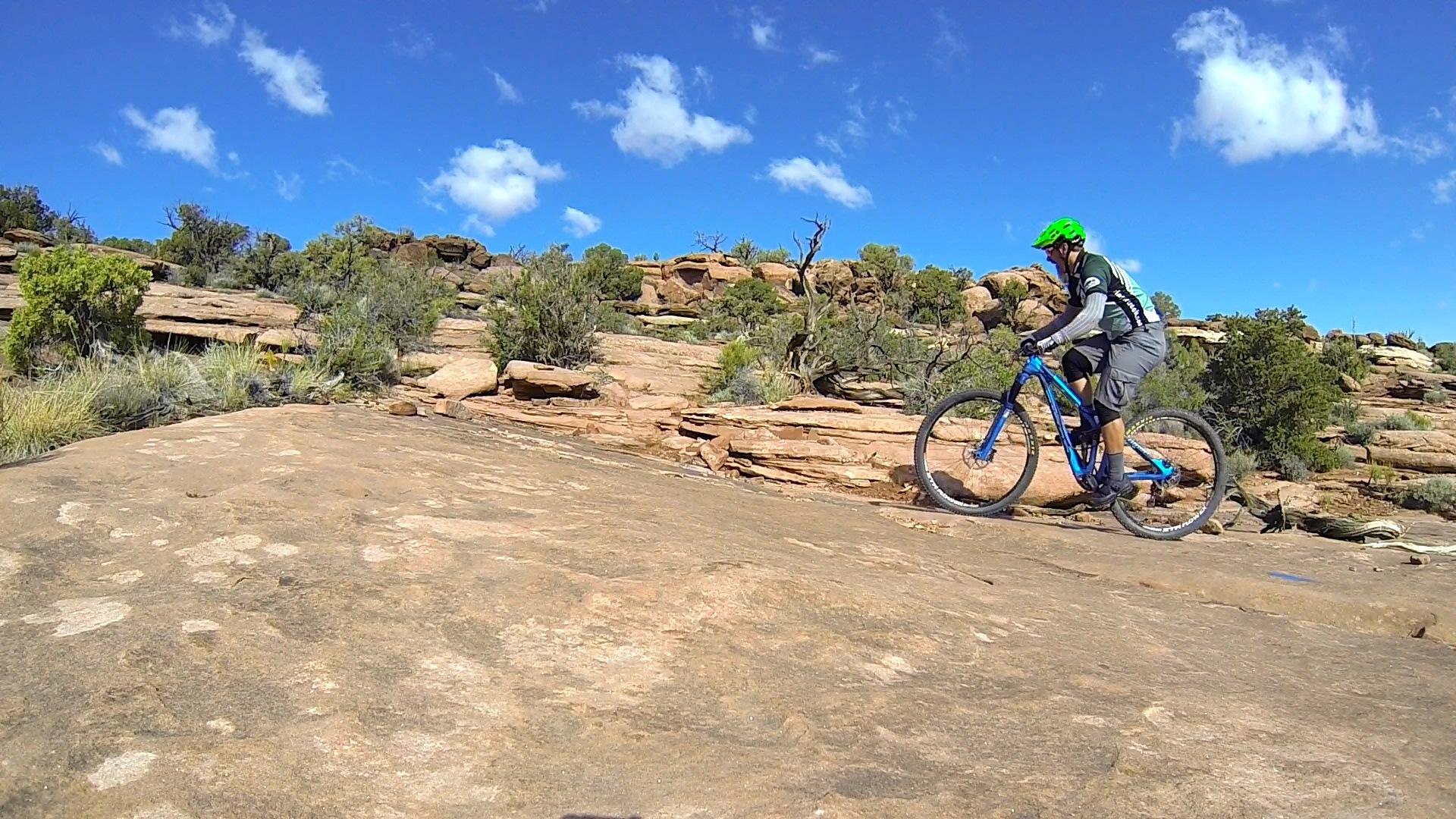 A mountain biker navigating rocky terrain under a clear blue sky, with sparse vegetation and rugged rock formations in the background. The biker is wearing a bright green helmet and is riding a blue bicycle, showcasing an adventurous outdoor setting. Captain Ahab mountain bike trail.