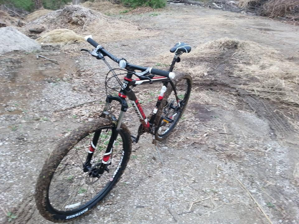 Trek X-Caliber 29: A red and black mountain bike is parked on a gravel path, with mud splattered on the tires, indicating recent use. Surrounding the bike are patches of dry grass and debris, suggesting a natural outdoor setting.