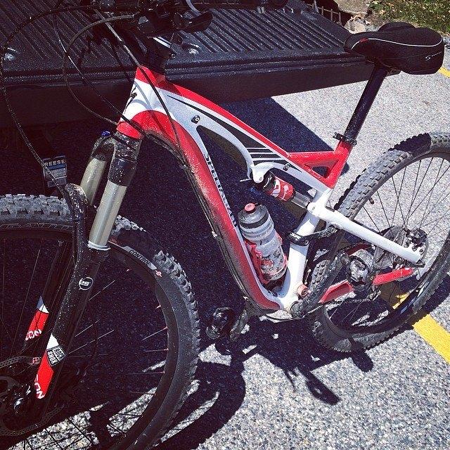 Specialized Camber: A red and white mountain bike parked on a gravel surface, with a water bottle attached to the frame. The bike features thick, knobby tires and a front suspension fork, indicating it is designed for off-road cycling. In the background, a truck bed can be partially seen.