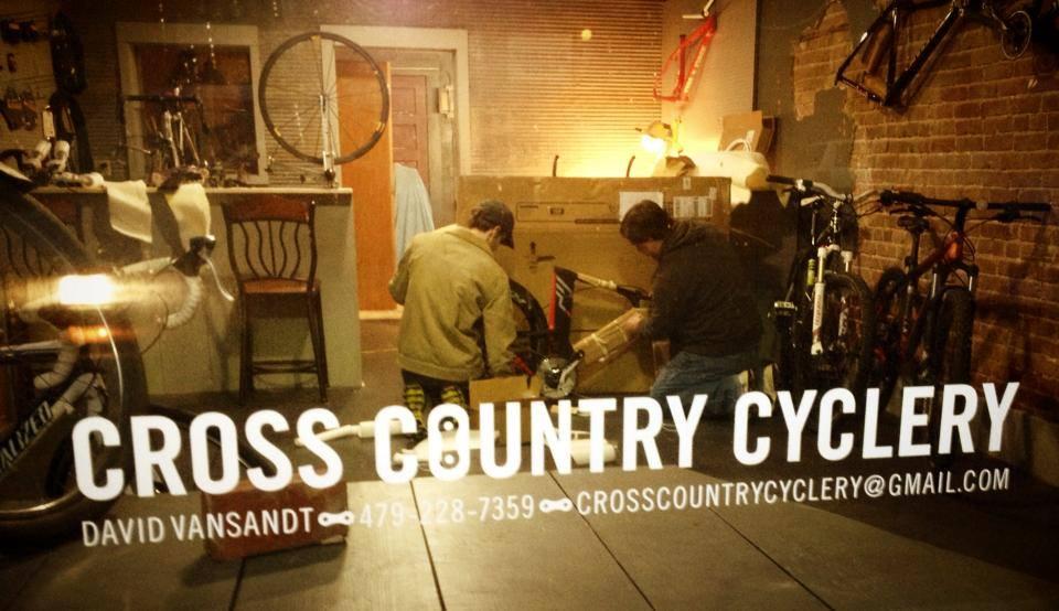 A dimly lit bicycle shop interior, featuring two individuals working on assembling or repairing bikes. In the foreground, the shop name "CROSS COUNTRY CYCLERY" is displayed prominently. The background includes various bicycles, tools, and an inviting workspace.
