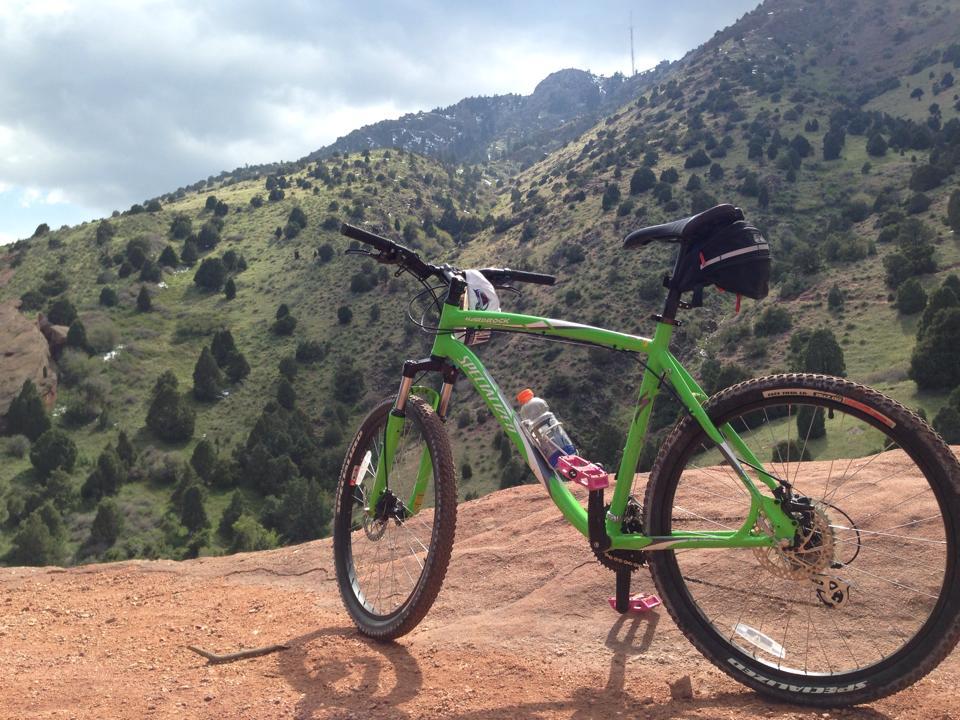 A green mountain bike rests on rocky terrain, overlooking a scenic landscape of rolling hills and trees under a cloudy sky. A water bottle is attached to the bike, ready for the journey ahead. Red Rocks / Dakota Ridge mountain bike trail.