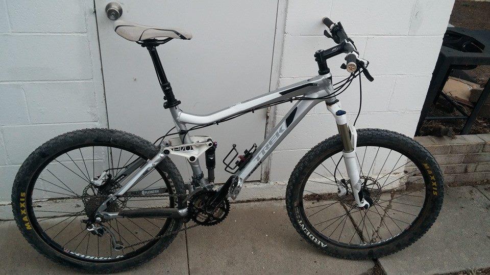 Trek Fuel EX 6: A silver and black mountain bike with thick tires and front suspension, parked against a white wall. The bike features a prominent brand name, gear components, and a detailed drivetrain, with a background showing a portion of a door and paved ground.