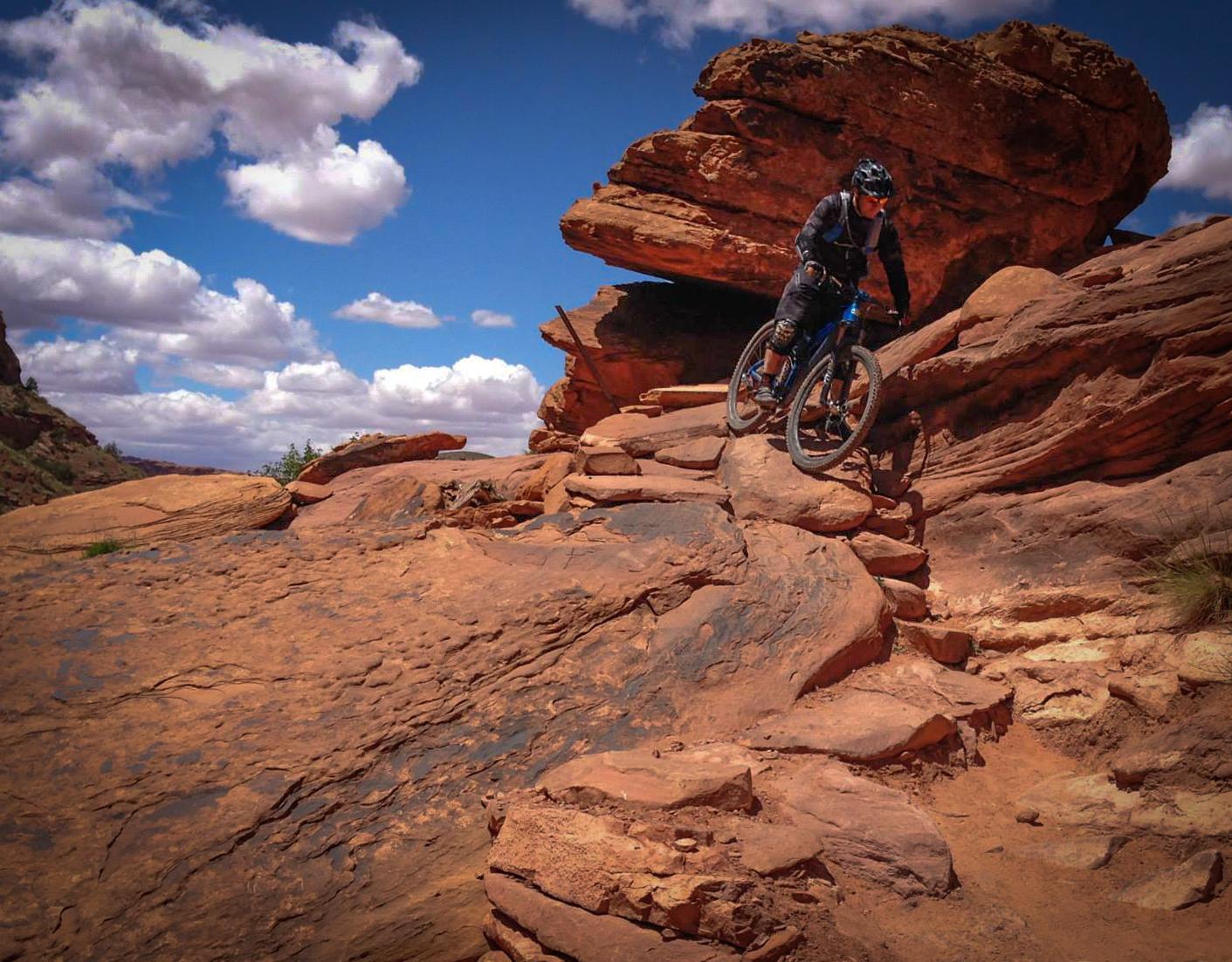 A mountain biker navigating a rocky terrain under a blue sky with fluffy clouds. The biker is positioned on a steep slope, skillfully riding over large stones and boulders in a rugged landscape. Porcupine Rim mountain bike trail.