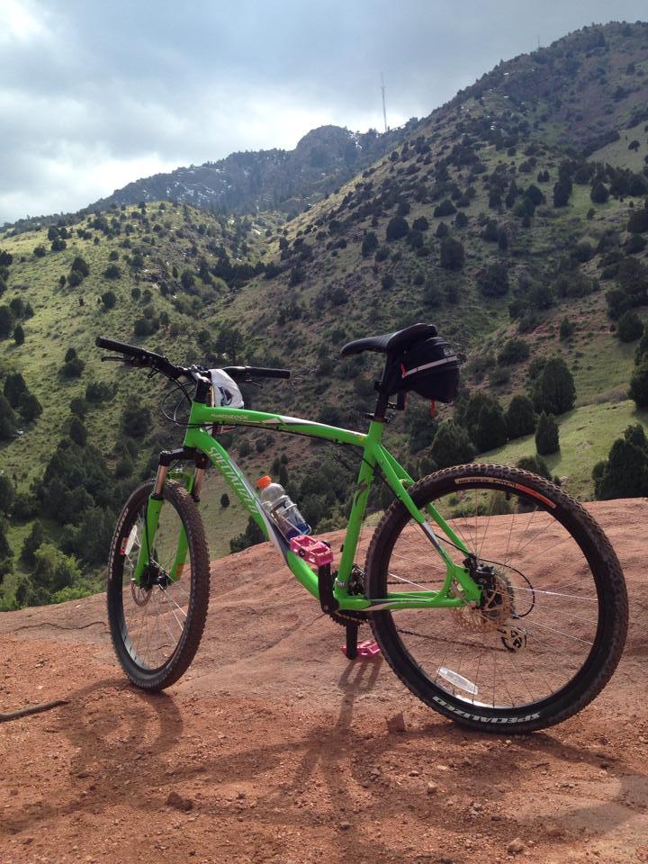A bright green mountain bike stands on rocky terrain with rolling hills and a cloudy sky in the background. A water bottle is mounted on the frame, and the landscape features a mix of greenery and rocky outcrops, hinting at a scenic outdoor location. Red Rocks / Dakota Ridge mountain bike trail.