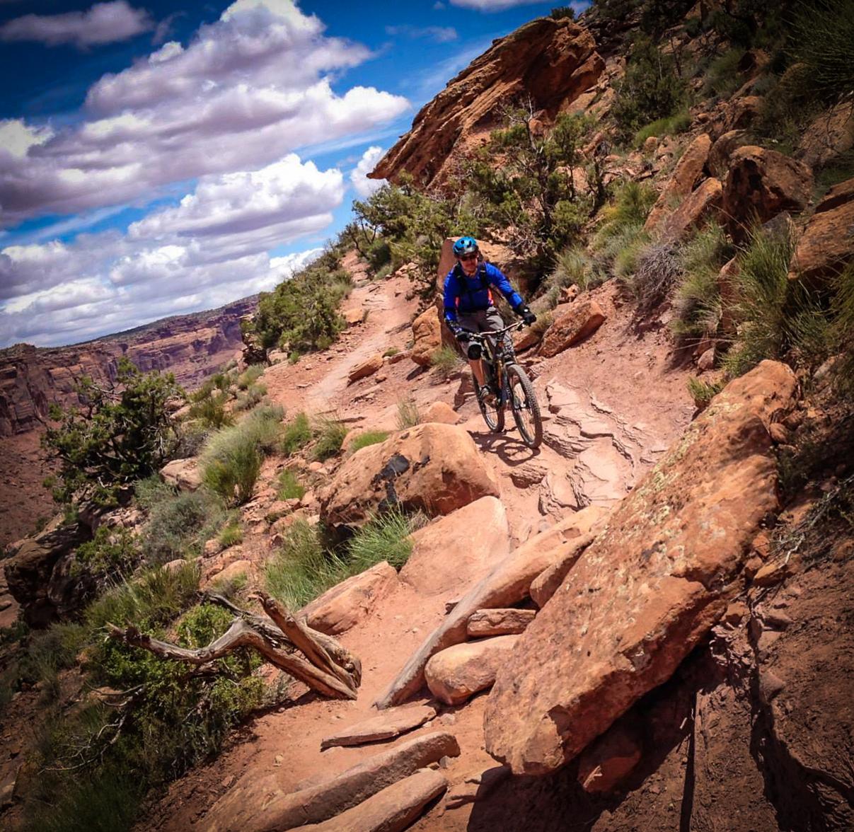 A mountain biker navigates a rocky trail in a desert landscape, surrounded by natural vegetation and dramatic rock formations under a partly cloudy sky. Porcupine Rim mountain bike trail.