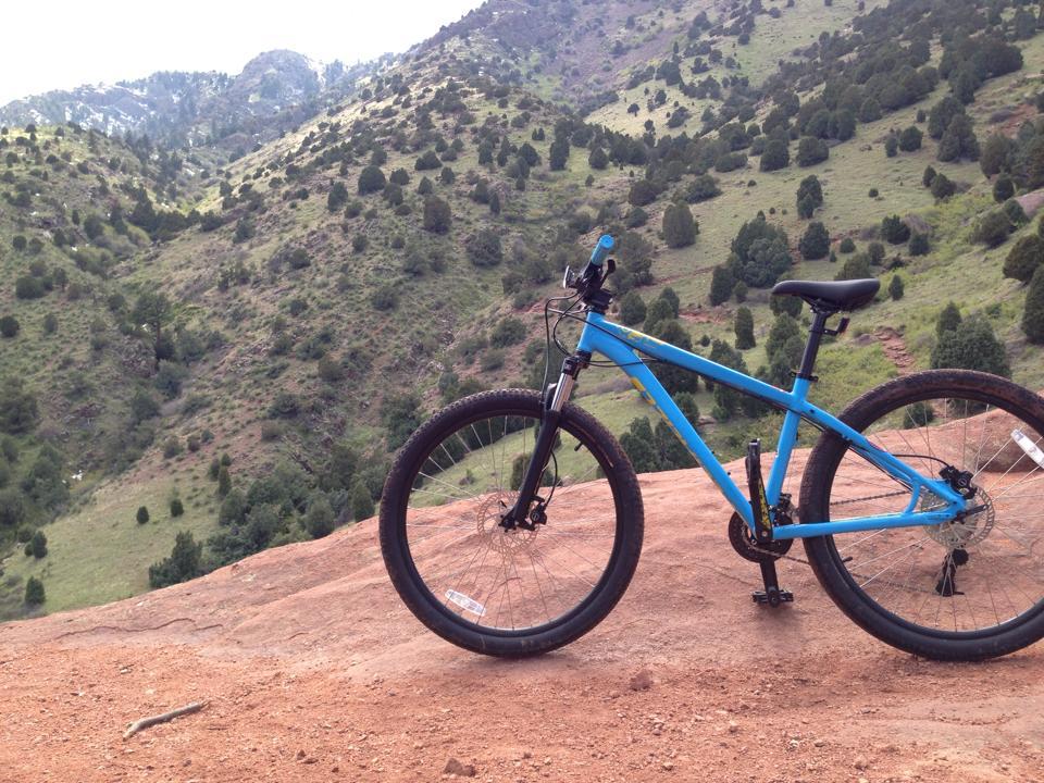 A blue mountain bike is positioned on a rocky ledge, overlooking a lush, green valley and rolling hills in the background. The scene captures a beautiful outdoor landscape with trees and rocky terrain. Red Rocks / Dakota Ridge mountain bike trail.
