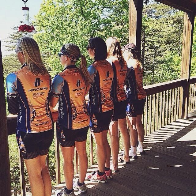 Five women in cycling jerseys are standing in a row on a wooden balcony, looking out at a green landscape. The jerseys are orange and blue, featuring the Hincapie brand. The scene is sunny, and the background includes trees and a clear sky.