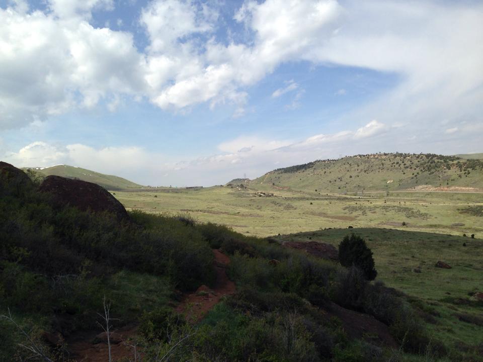 A panoramic view of a lush green landscape featuring rolling hills, rocky outcrops, and a partly cloudy sky. The foreground includes shrubs and a dirt path leading into the distance, while the horizon showcases a gentle slope of hills under a blue sky. Red Rocks / Dakota Ridge mountain bike trail.