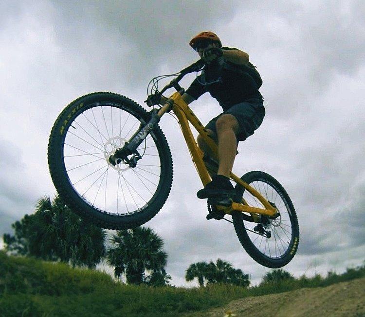 Yeti SB-75: A person riding a yellow mountain bike performs a jump over a dirt ramp against a cloudy sky, with trees visible in the background.