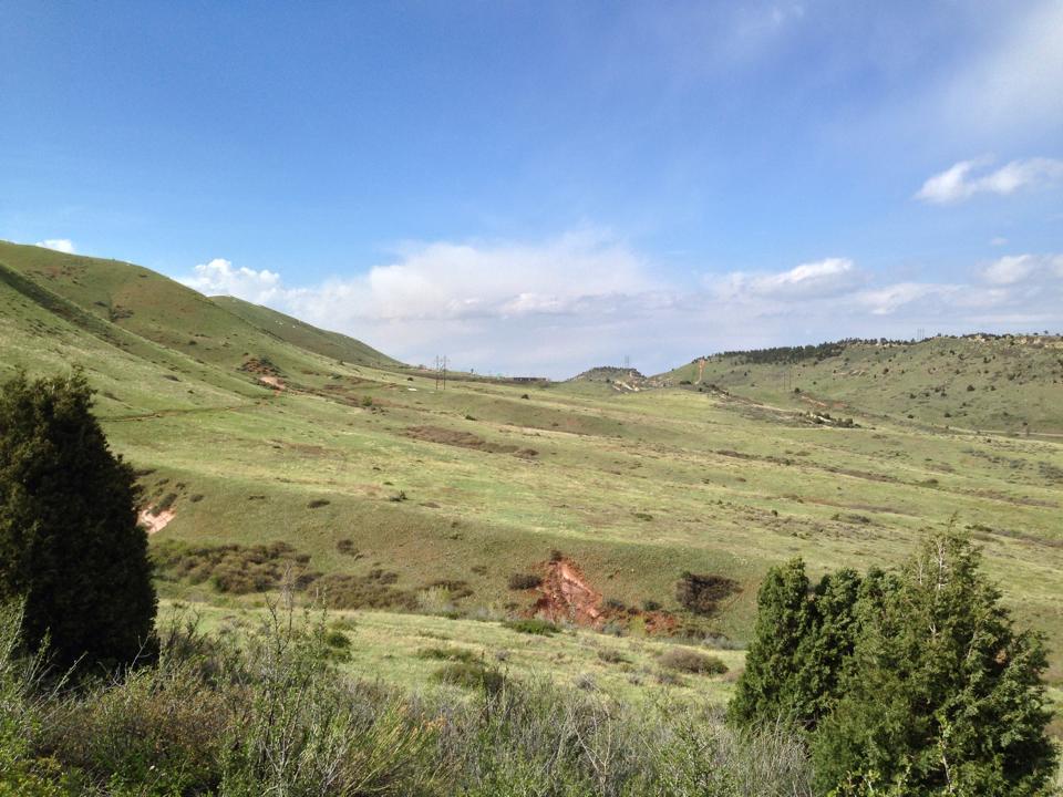 A panoramic view of rolling green hills under a bright blue sky with scattered clouds. In the foreground, there are patches of shrubs and trees, while the background features more hills and a distant horizon with a few power lines. The scene conveys a sense of tranquility and open space in a natural landscape. Red Rocks / Dakota Ridge mountain bike trail.