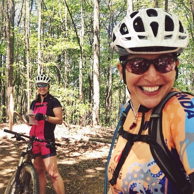 Two women in athletic cycling gear are standing on a wooded trail during daytime. One woman, wearing a colorful jersey and a white helmet, is smiling at the camera, while the other woman, dressed in a pink outfit, is holding a beverage and standing next to her mountain bike. Lush green trees surround them, creating a vibrant outdoor atmosphere.