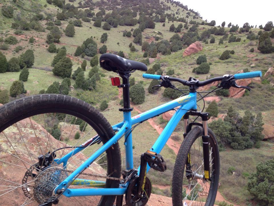 A blue mountain bike parked on a rocky outcrop with a scenic view of rolling green hills and sparse trees in the background. Red Rocks / Dakota Ridge mountain bike trail.