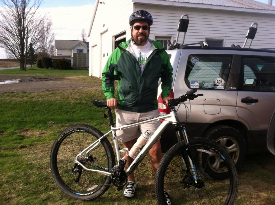 Raleigh Talus: A man wearing a helmet and a green jacket stands next to a mountain bike, smiling at the camera. The background features a grassy area, a parked vehicle, and a building. The man is dressed in shorts and sandals, indicating a casual outdoor setting.