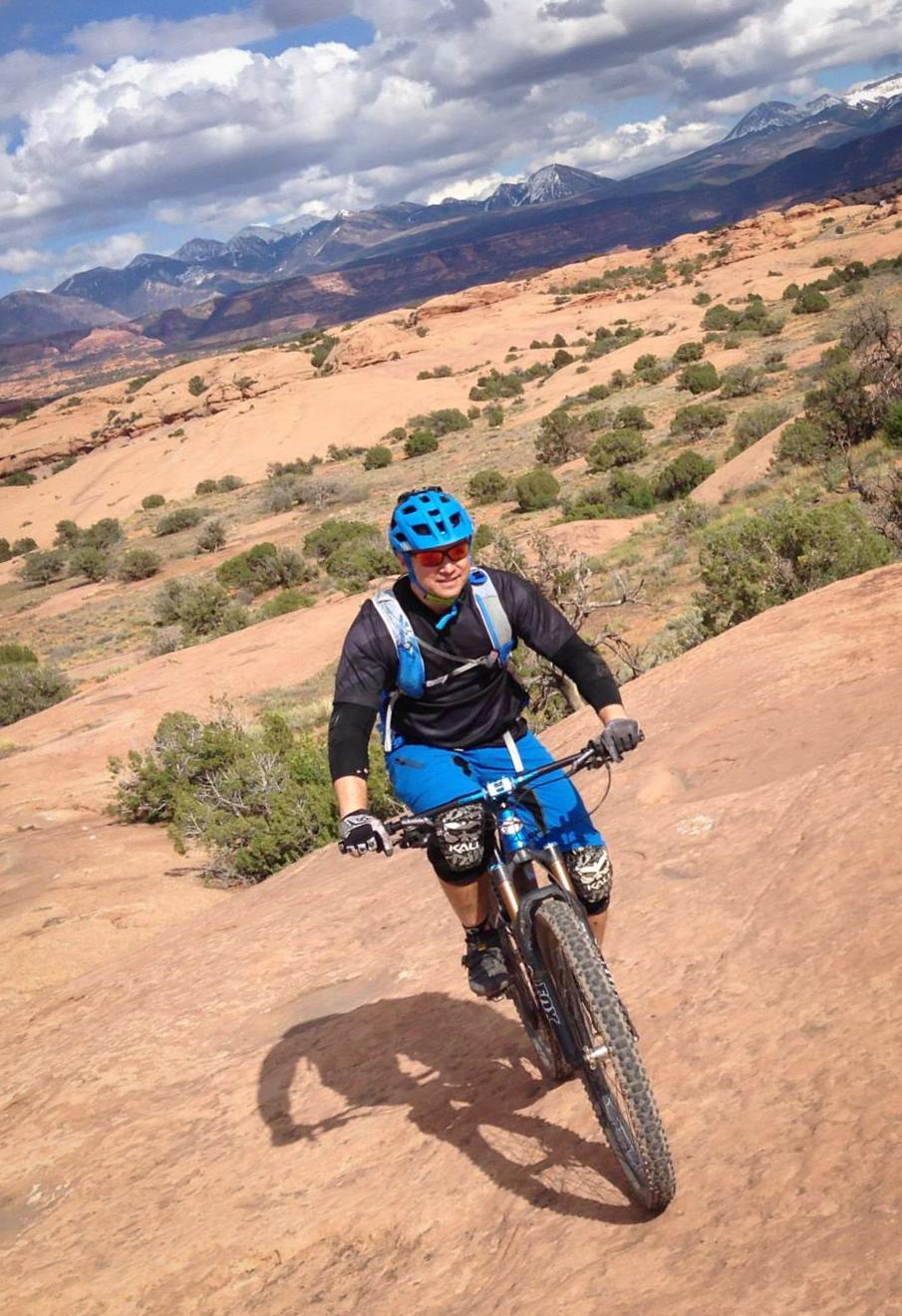 A mountain biker riding on rocky terrain with a scenic backdrop of mountains and a partly cloudy sky. The rider is wearing a blue helmet, sunglasses, and protective gear, while navigating a slope with desert vegetation nearby. Slickrock mountain bike trail.