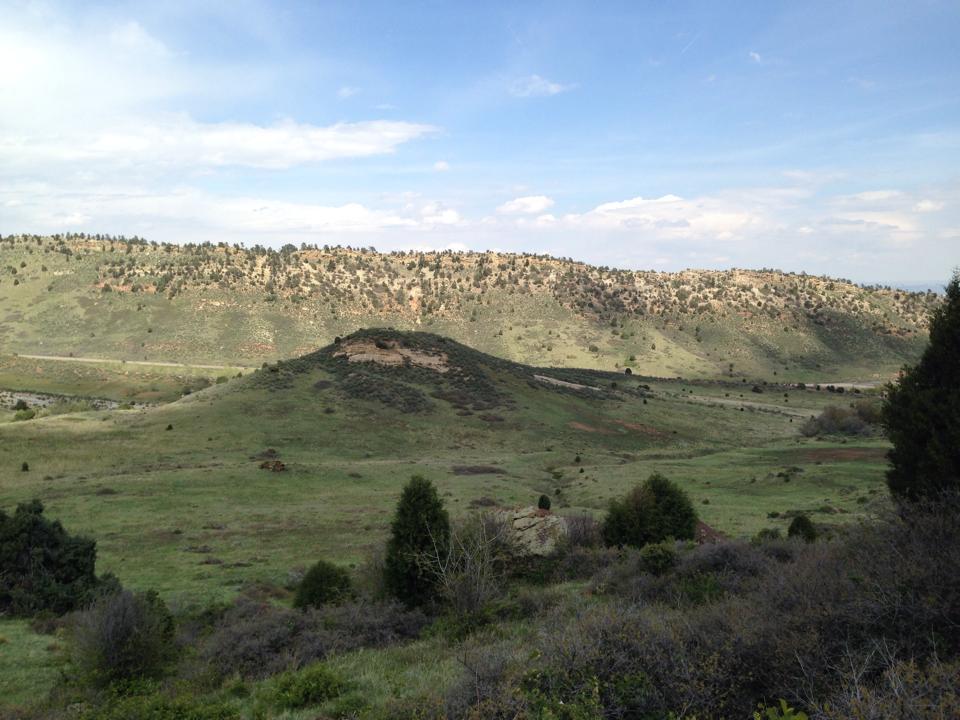 A panoramic view of rolling green hills under a partly cloudy sky. The landscape features a variety of vegetation, including low shrubs and scattered trees, with a visible road winding through the valley in the background. Red Rocks / Dakota Ridge mountain bike trail.
